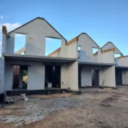 Wznoszenie segmentu domu z białych bloczk&oacute;w, widoczne otwory okienne i drzwiowe, szalunki stropowe, oraz narzędzia budowlane na placu budowy.