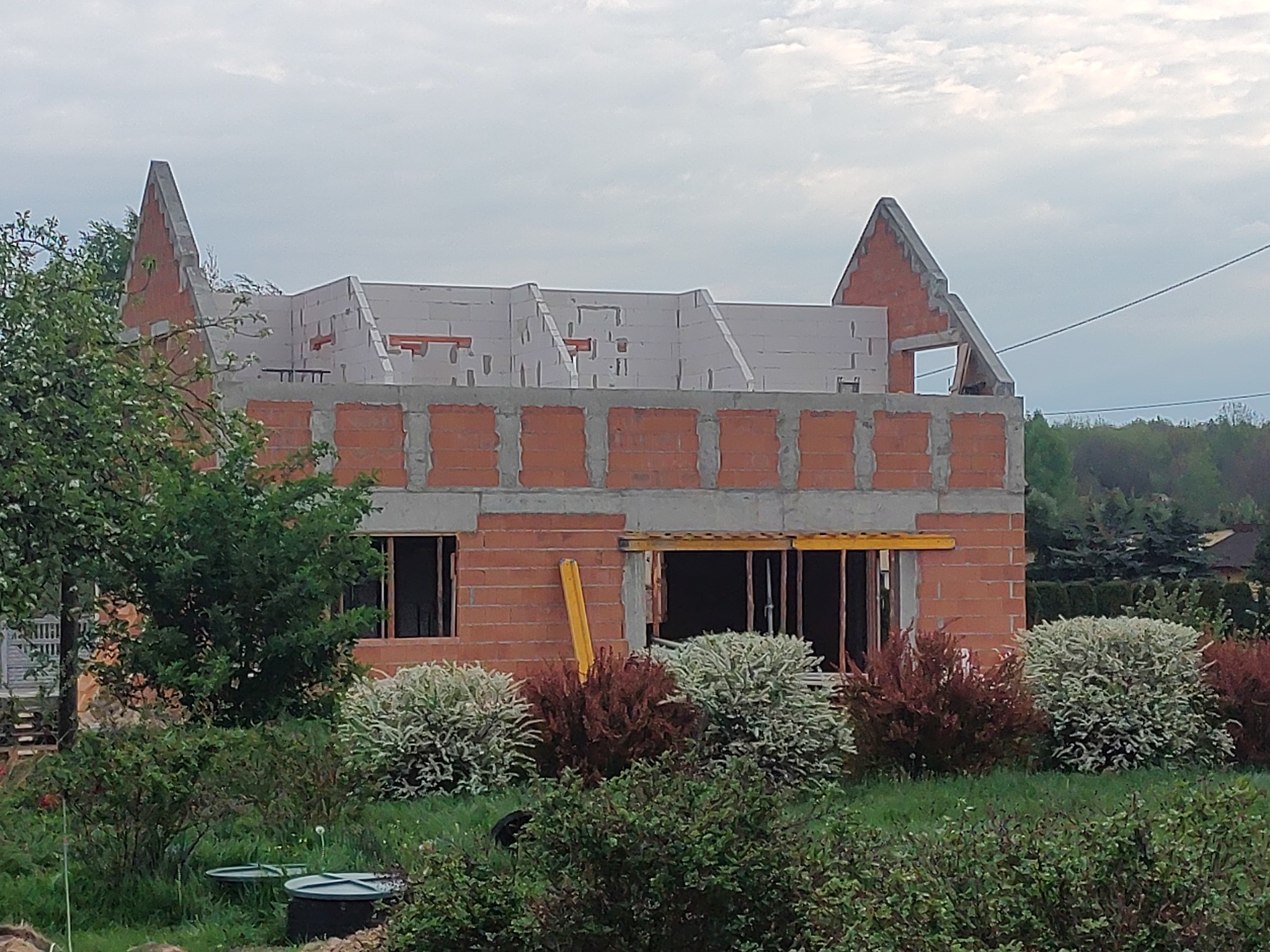 Dom w trakcie budowy: czerwona cegła, białe bloczki, widoczne otwory okienne, żółta poziomica. W tle zieleń i pochmurne niebo.