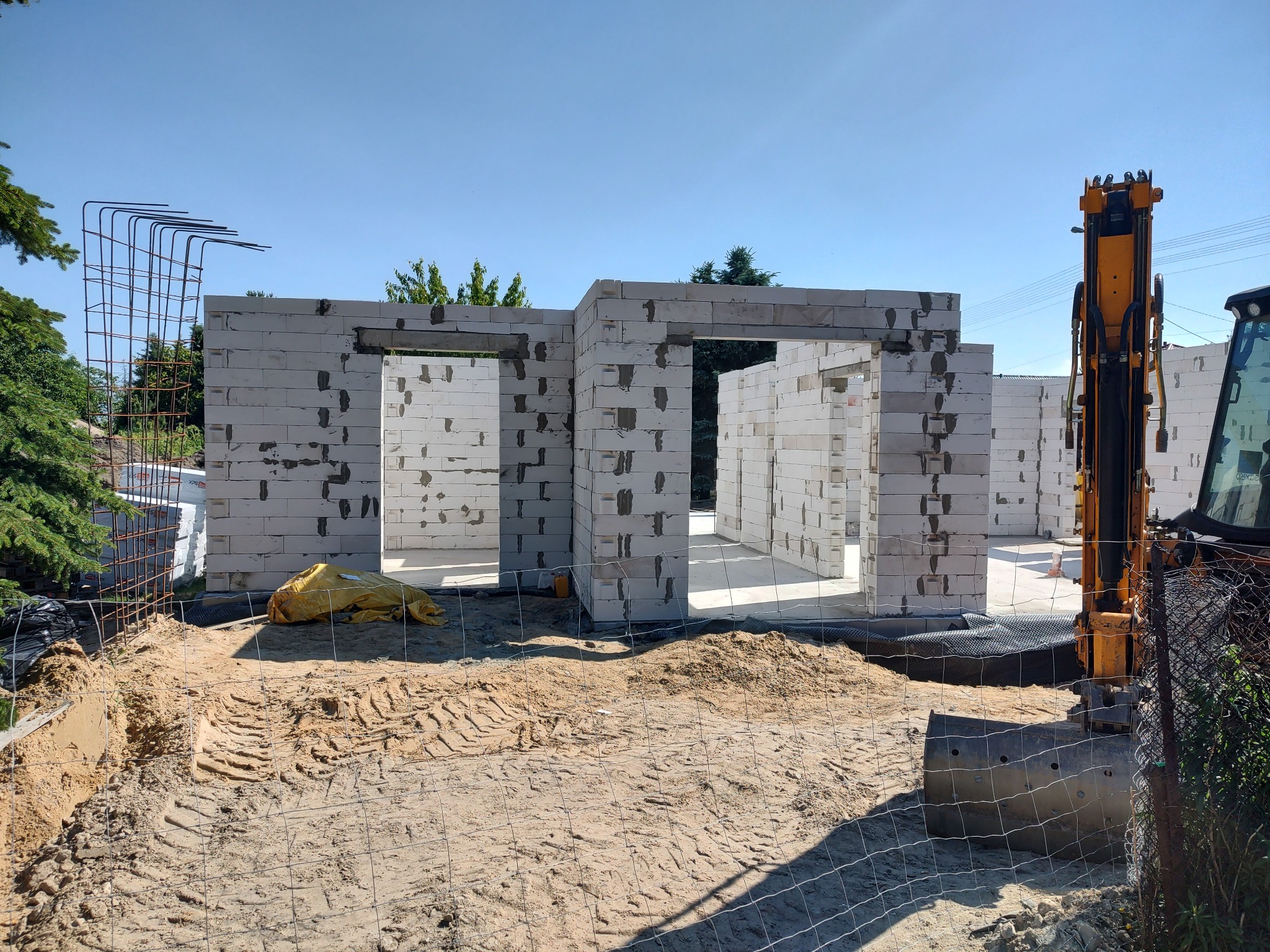 Ściany domu z betonu komórkowego w trakcie murowania, widoczny szalunek, zbrojenie i koparka na działce. Budowa domu w toku.