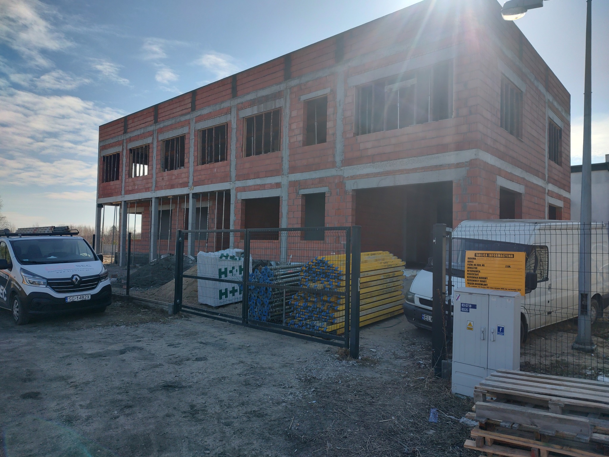 Budynek dwukondygnacyjny w trakcie budowy z czerwonej cegły, widoczny przez metalową bramę. Na pierwszym planie materiały budowlane i samochód dostawczy.