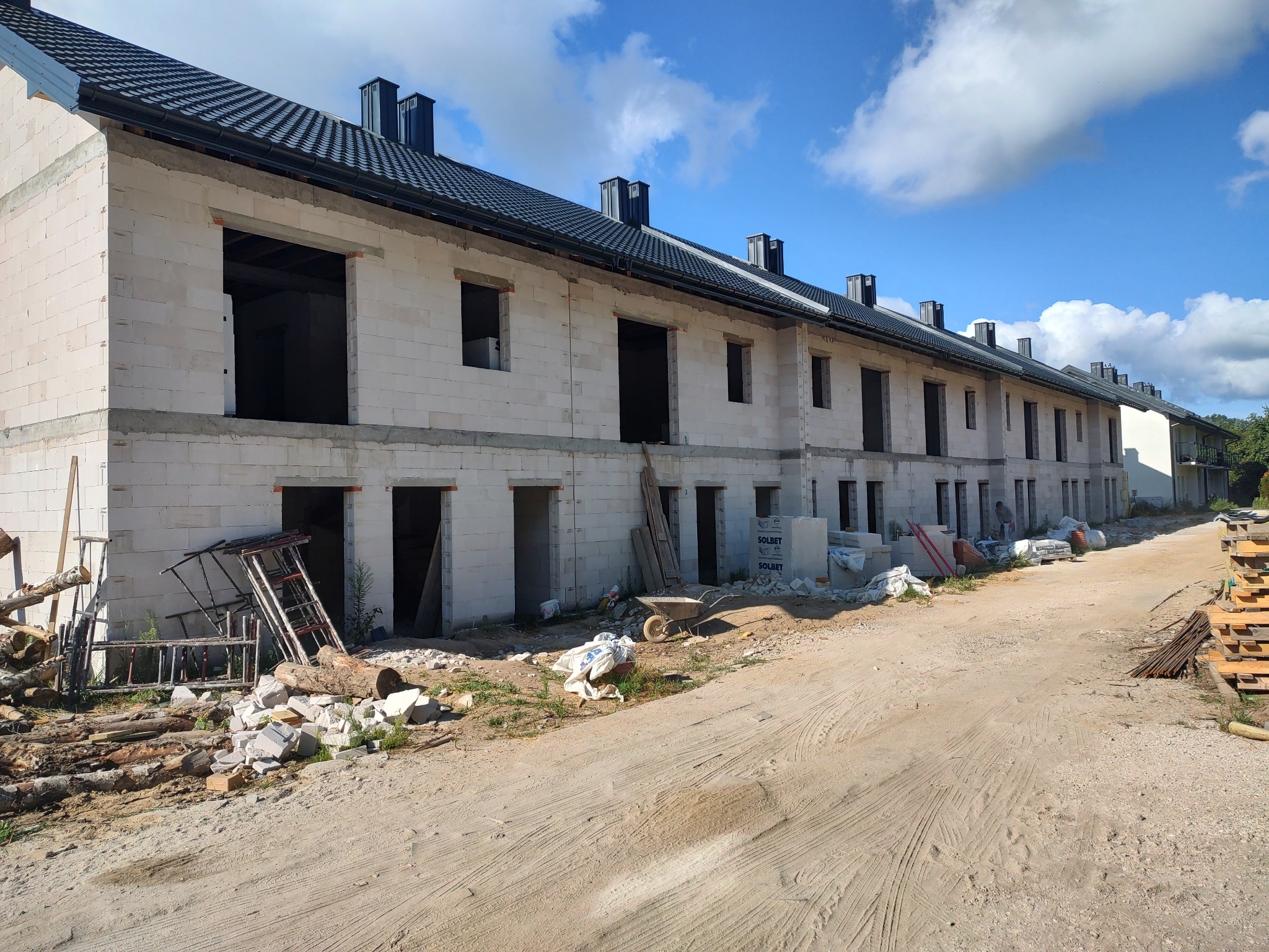 Wznoszenie szeregu domów z białych bloczków, z widocznymi otworami okiennymi i drzwiowymi. Na pierwszym planie materiały budowlane i narzędzia.