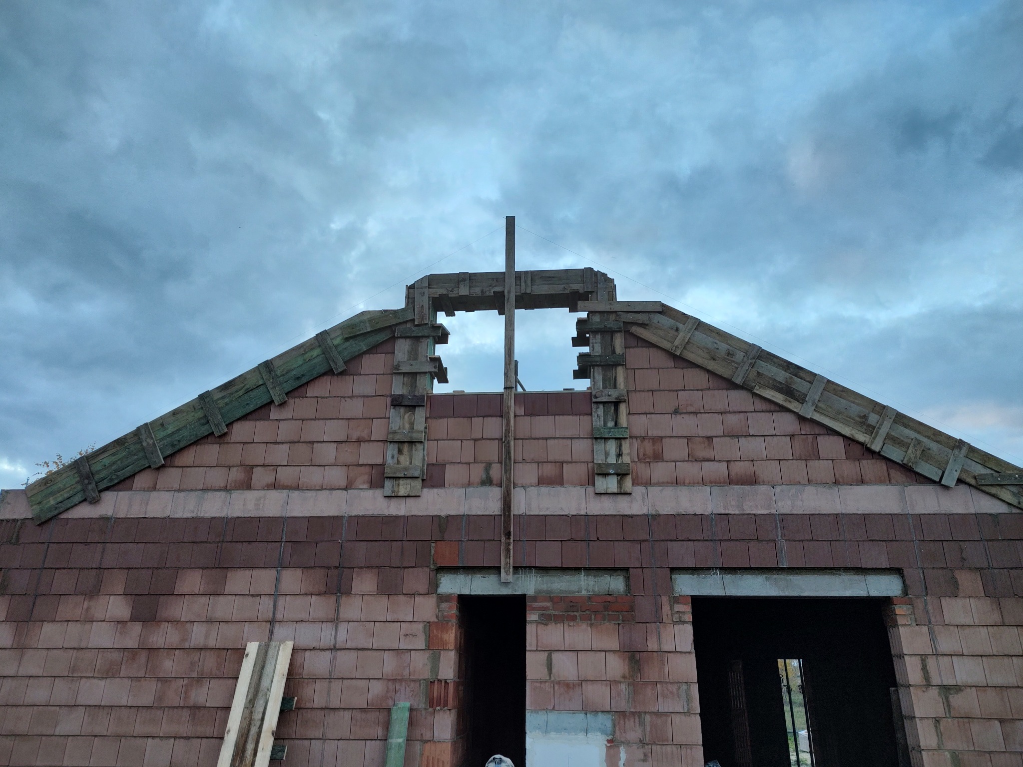 Wznoszenie domu z keramzytu: widok na fragment ściany szczytowej z otworami okiennymi w trakcie murowania, z drewnianym szalunkiem i pochmurnym niebem w tle.