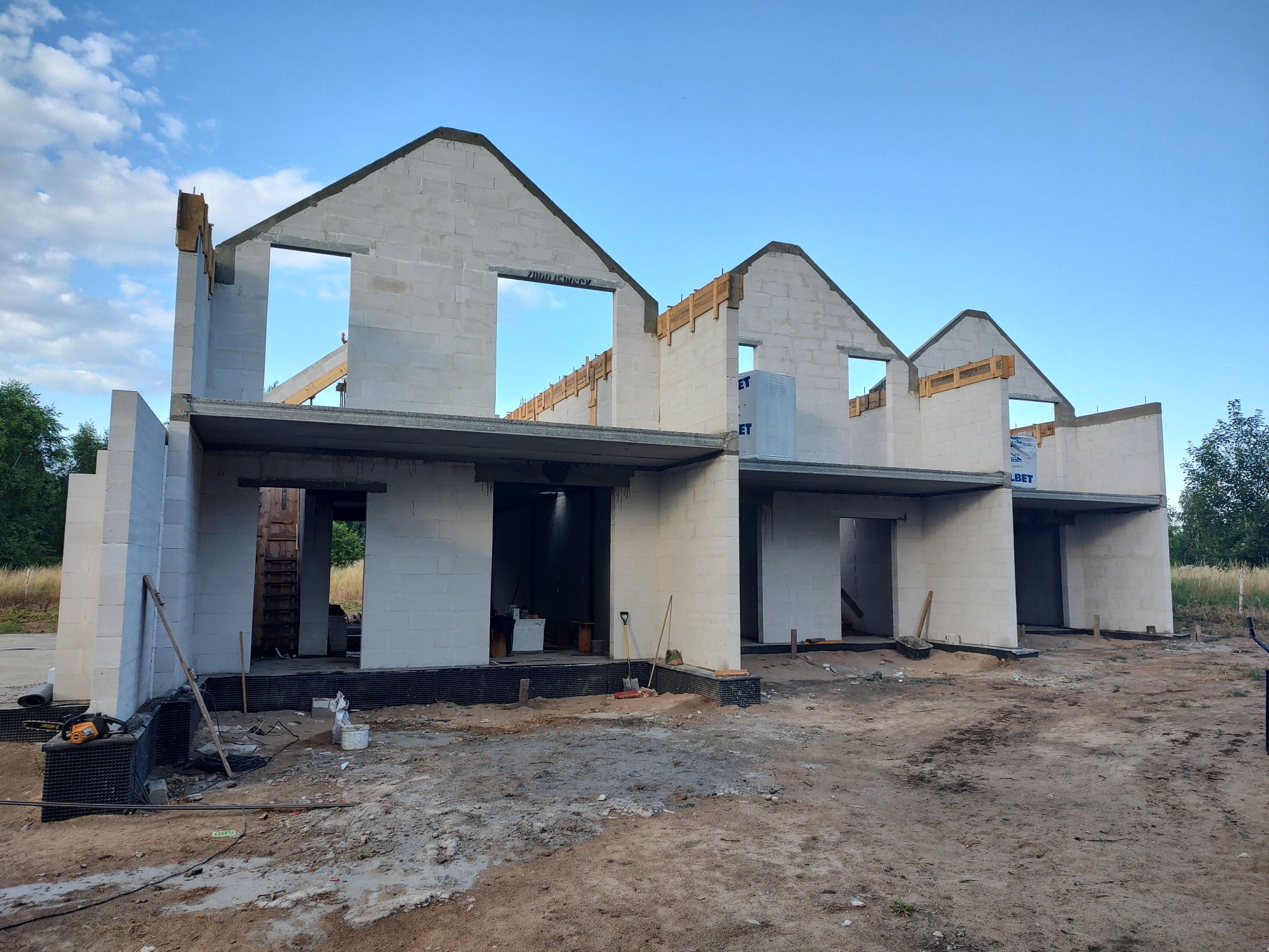 Wznoszenie segmentu domu z białych bloczków, widoczne otwory okienne i drzwiowe, szalunki stropowe, oraz narzędzia budowlane na placu budowy.