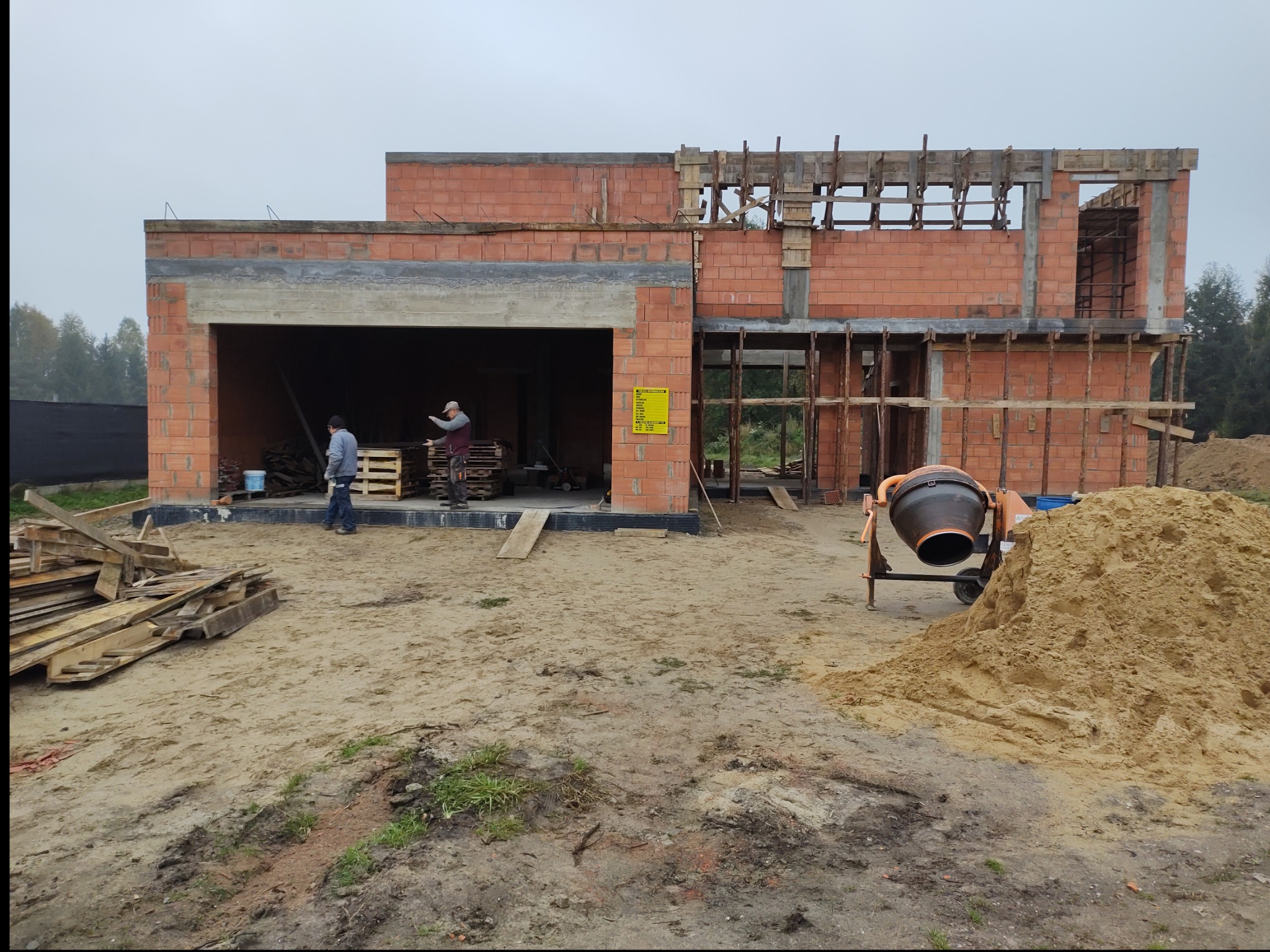 Budowa domu z czerwonej cegły: widok na dwukondygnacyjny budynek w trakcie murowania ścian, z widoczną betoniarką i pracownikami na placu budowy.