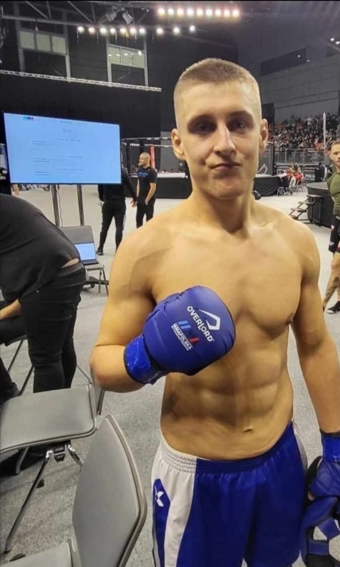 Młody sportowiec MMA z niebieskimi rękawicami Overlord, prezentuje wyrzeźbioną sylwetkę na tle trybun podczas zawodów.