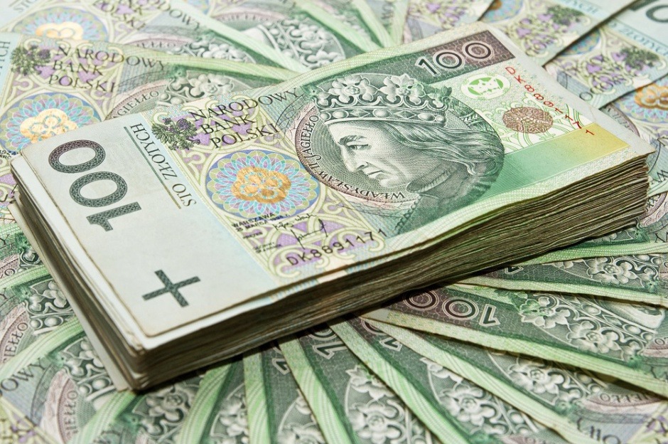 Stos banknotów 100 zł na tle rozłożonych pieniędzy. Widoczny portret Władysława Jagiełły i nominał. Delikatne światło podkreśla detale banknotów.