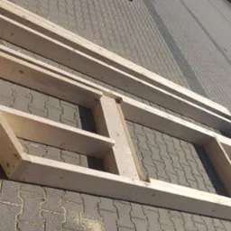 Drewniana rama konstrukcyjna z jasnego drewna na tle szarej kostki brukowej, widok z g&oacute;ry pod kątem. Elementy połączone w regularne prostokąty.