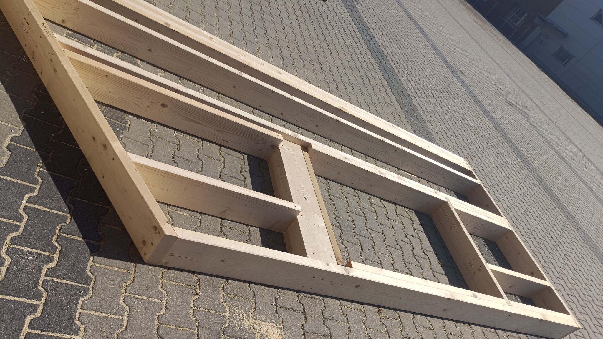 Drewniana rama konstrukcyjna z jasnego drewna na tle szarej kostki brukowej, widok z góry pod kątem. Elementy połączone w regularne prostokąty.