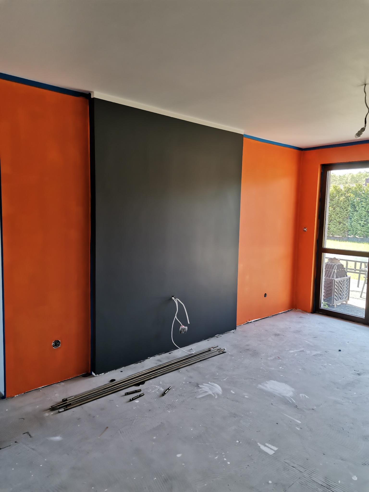 Wnętrze w trakcie malowania: pomarańczowe ściany zabezpieczone taśmą, czarny fragment ściany, rury na podłodze, widoczne gniazdko elektryczne i przewody.