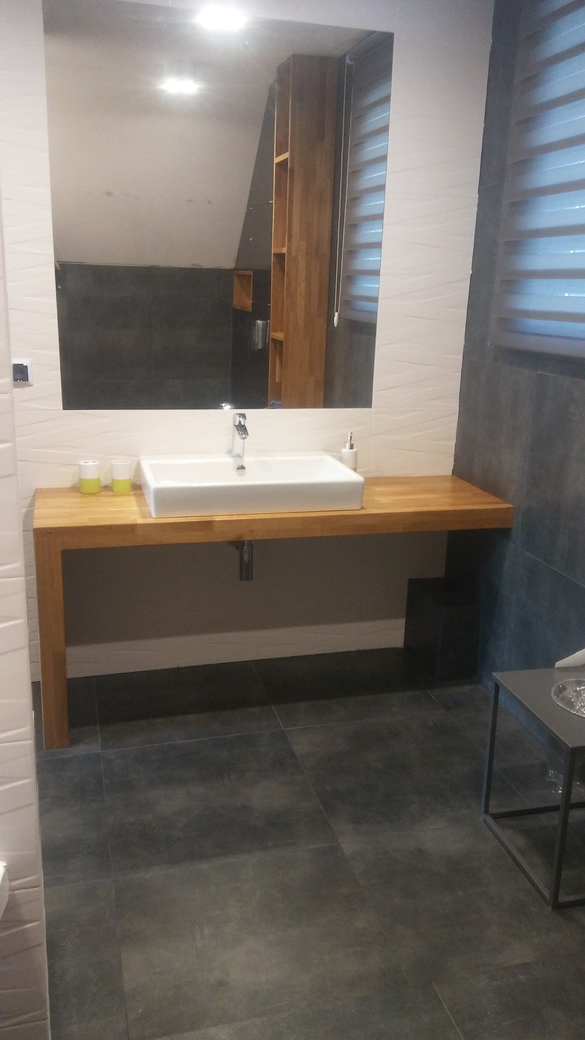 Nowoczesna łazienka z drewnianym blatem pod umywalką, duże lustro z drewnianą półką, szare płytki na podłodze i ścianach, minimalistyczny design.