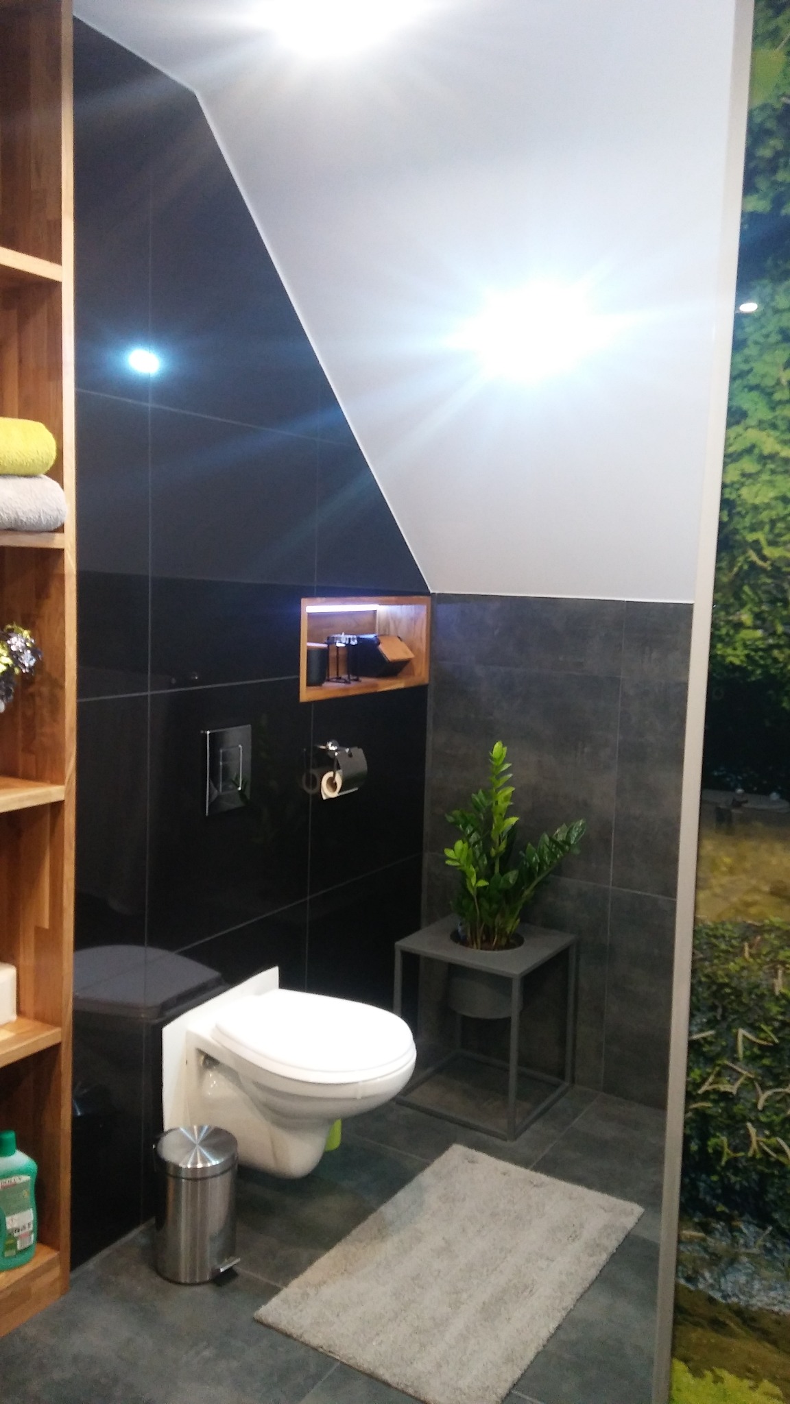 Nowoczesna łazienka z czarnymi płytkami, podświetlaną wnęką i rośliną w minimalistycznej donicy. Uwagę przykuwa skośny sufit i drewniany regał.