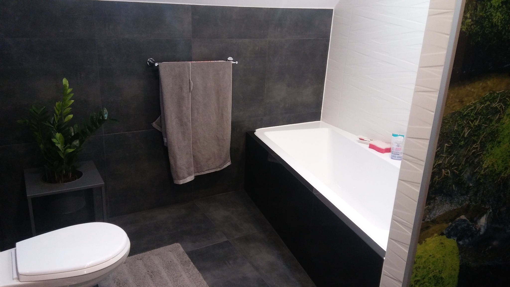 Nowoczesna łazienka z ciemnymi płytkami, białą wanną i toaletą. Na ścianie wisi ręcznik, a obok toalety stoi roślina doniczkowa. Minimalistyczny design.