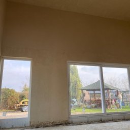 R4-BUD OLEH RUDENKO - Ściana po nałożeniu gładzi, widoczne duże okna z widokiem na ogród z altaną. Ściana w odcieniach beżu, surowy stan podłogi. Praca w trakcie wykończenia.