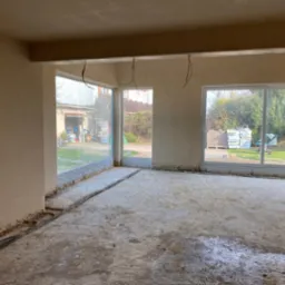 Surowy stan wnętrza po tynkowaniu ścian, z widocznymi kablami w podłodze i dużymi oknami z widokiem na zewnątrz. W tle garaż i ułożone materiały budowlane.