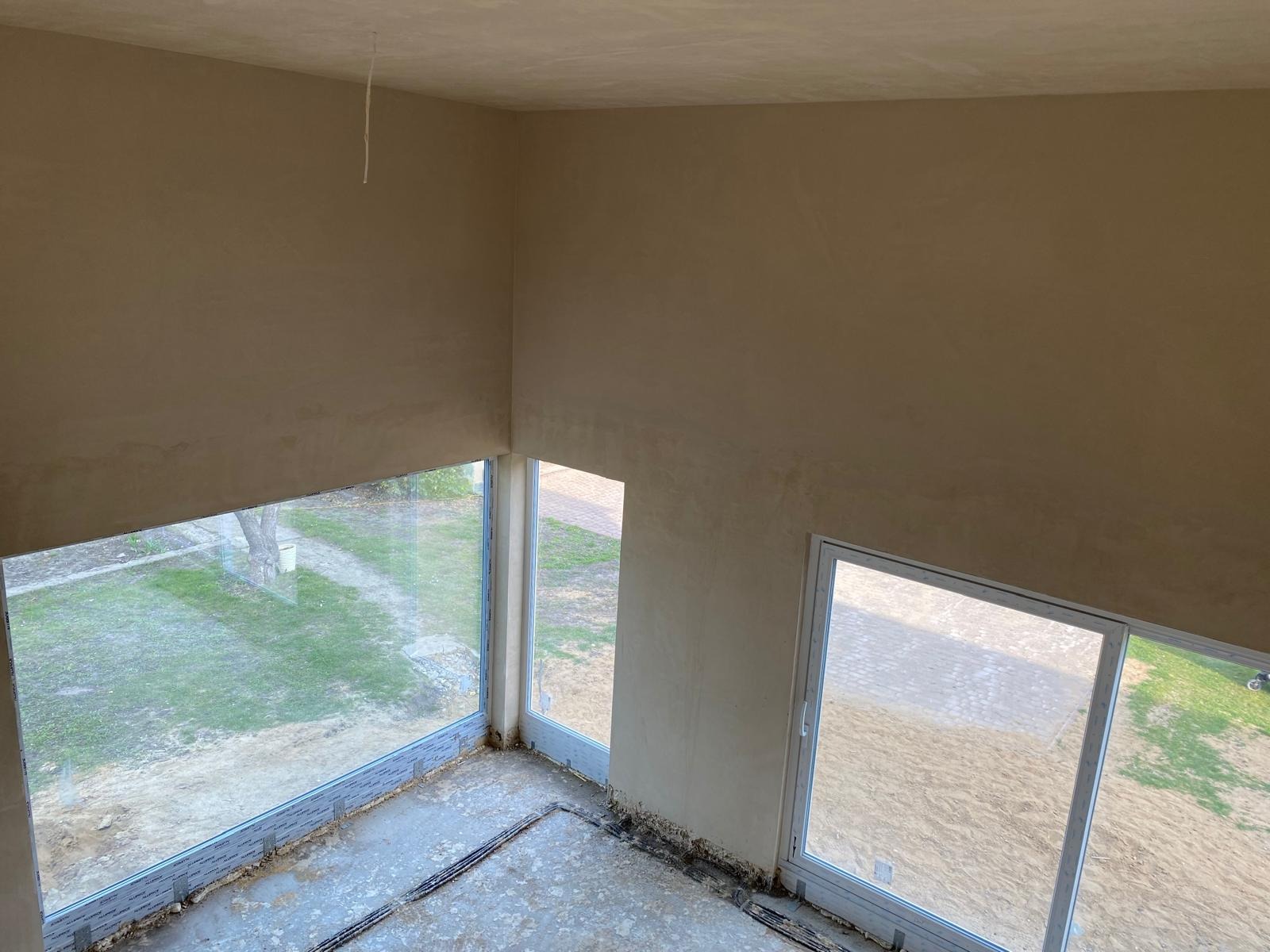 Wnętrze po nałożeniu tynku, widok z góry. Ściany o jednolitej fakturze, duże okna z widokiem na zewnątrz. Widoczne kable na podłodze i surowy stan wykończenia.