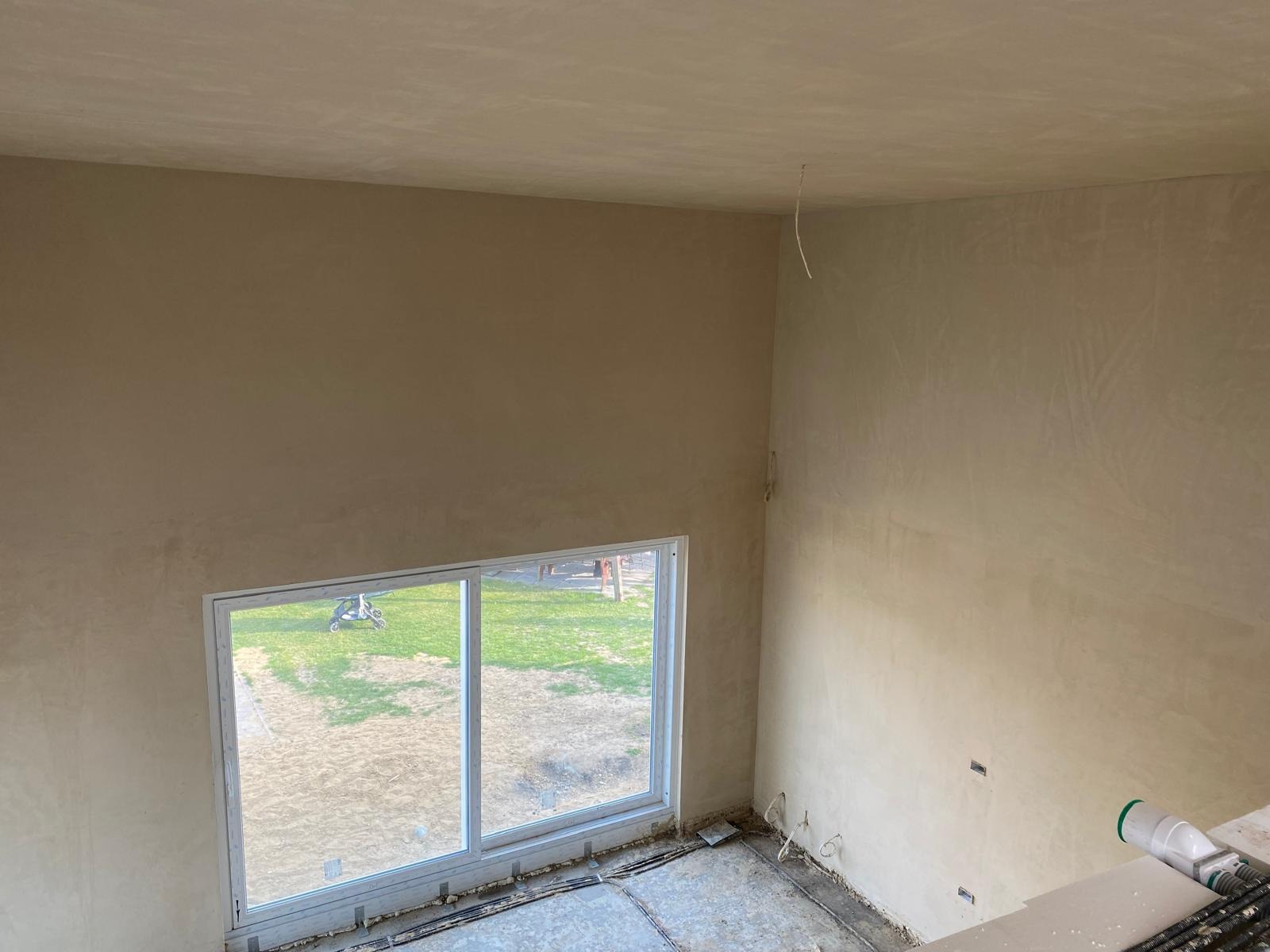 Wysokie wnętrze po gipsowaniu. Ściany z fakturą w odcieniach beżu, duże okno z widokiem na zieleń. Kabel zwisa z sufitu. Podłoga surowa, widać instalacje.