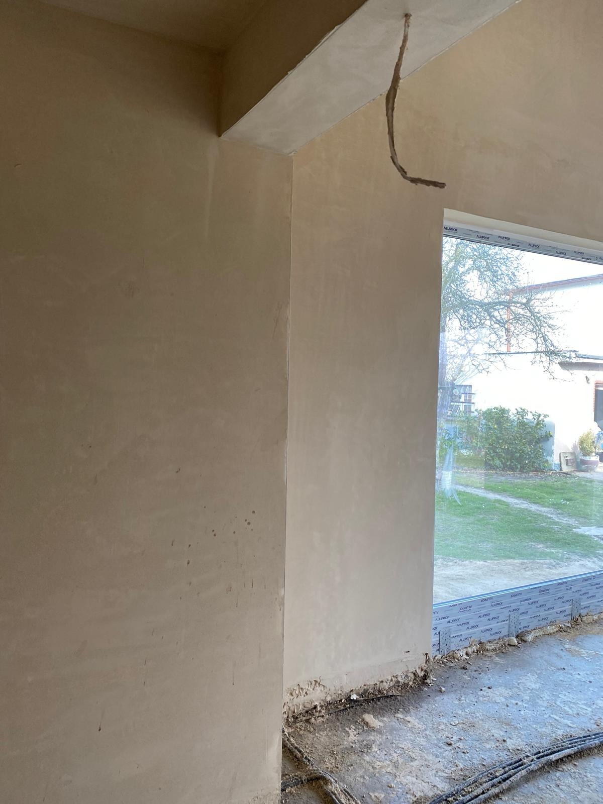 Ściana po tynkowaniu maszynowym, widoczne przewody elektryczne wystające z sufitu, okno z widokiem na zewnątrz.