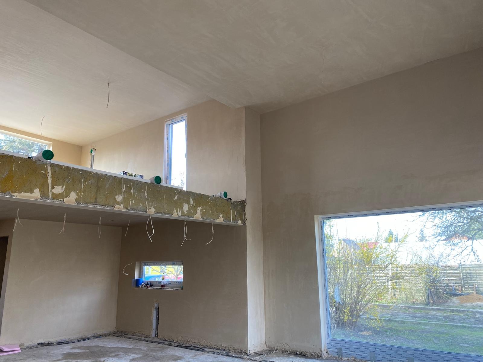 Wnętrze po szpachlowaniu: jasne ściany, okna z widokiem na zieleń, widoczne elementy konstrukcyjne stropu. Gładkie powierzchnie przygotowane do dalszych prac wykończeniowych.