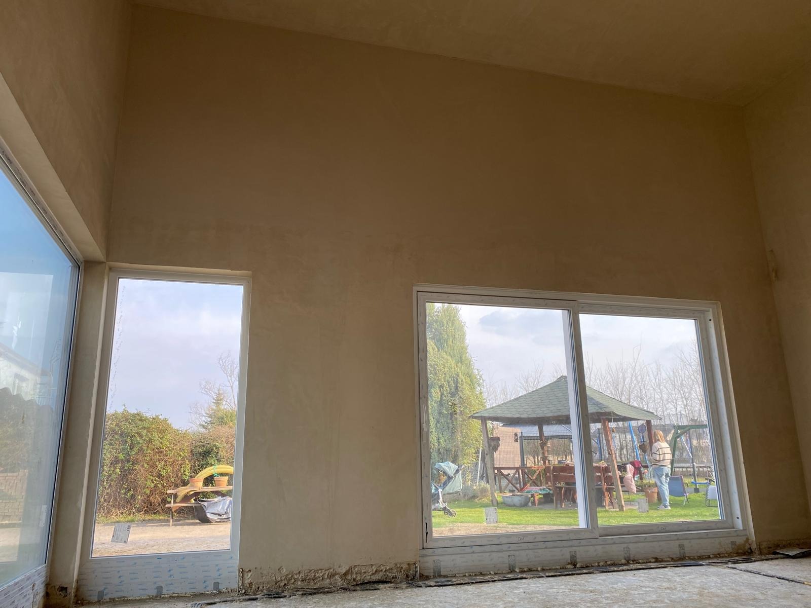 Ściana po nałożeniu gładzi, widoczne duże okna z widokiem na ogród z altaną. Ściana w odcieniach beżu, surowy stan podłogi. Praca w trakcie wykończenia.