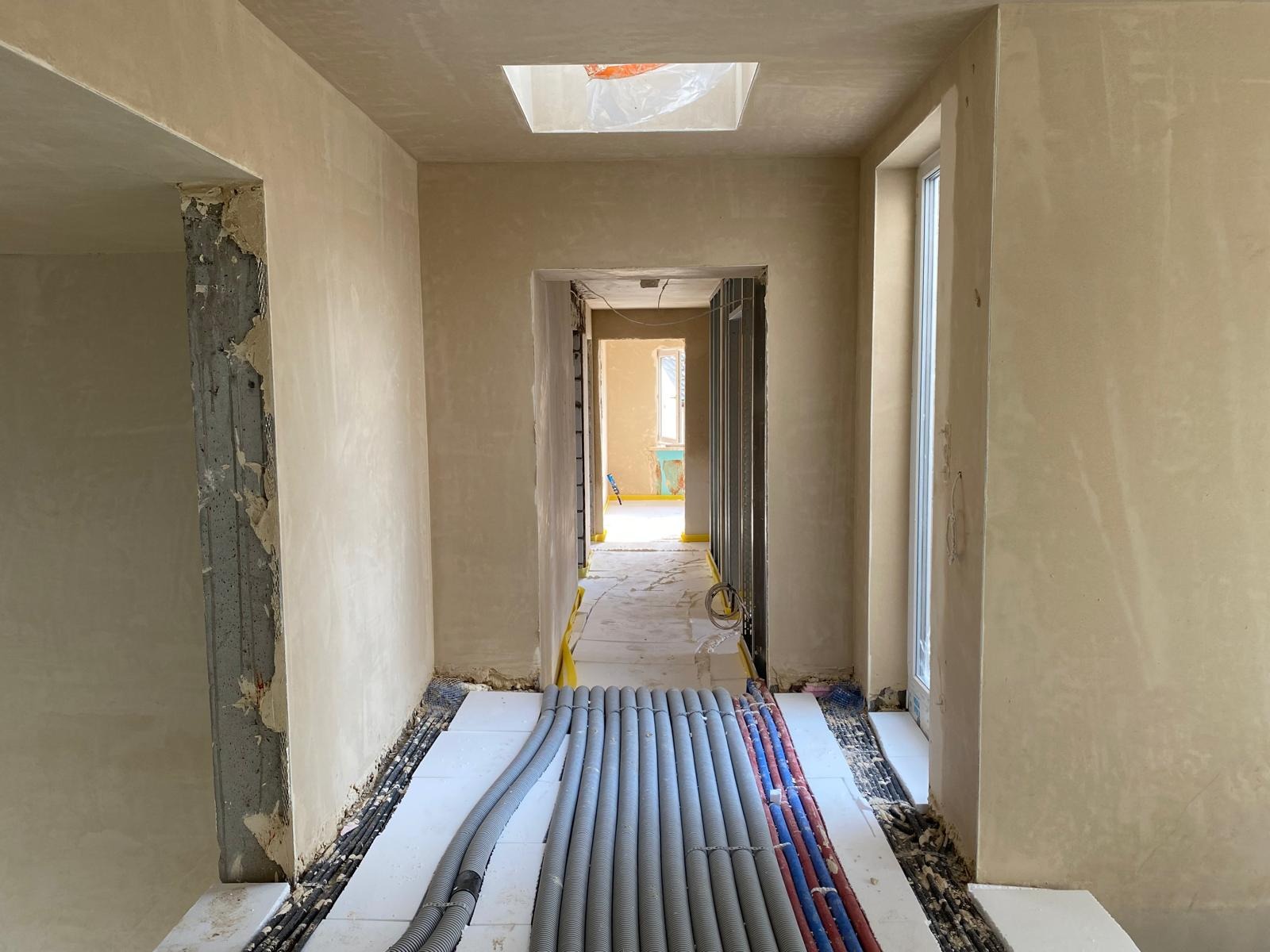 Korytarz w trakcie prac wykończeniowych: ściany pokryte tynkiem, widoczne rury instalacji podłogowej na izolacji, sufit z oknem dachowym. W tle widok na kolejne pomieszczenia.