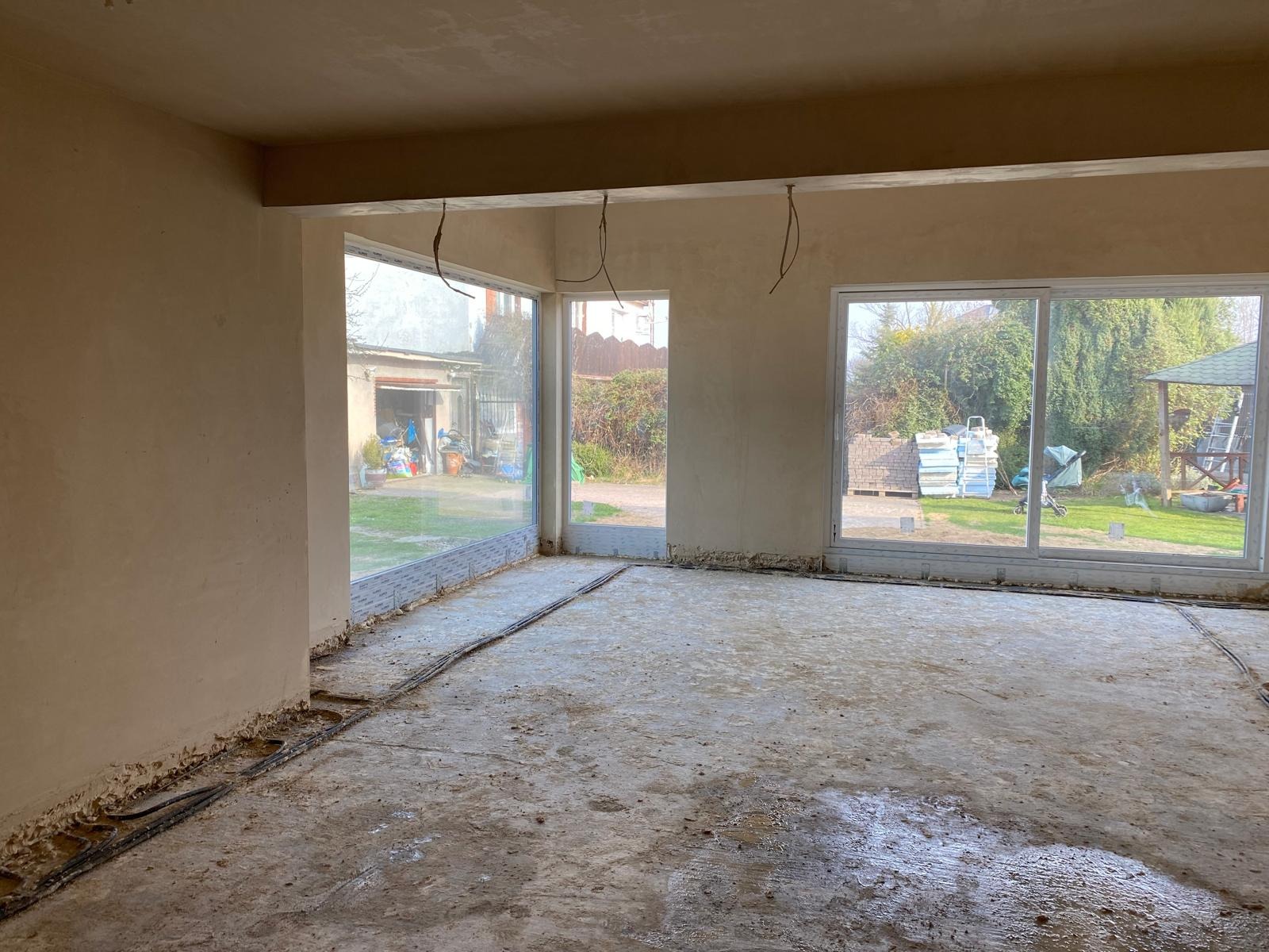 Surowy stan wnętrza po tynkowaniu ścian, z widocznymi kablami w podłodze i dużymi oknami z widokiem na zewnątrz. W tle garaż i ułożone materiały budowlane.