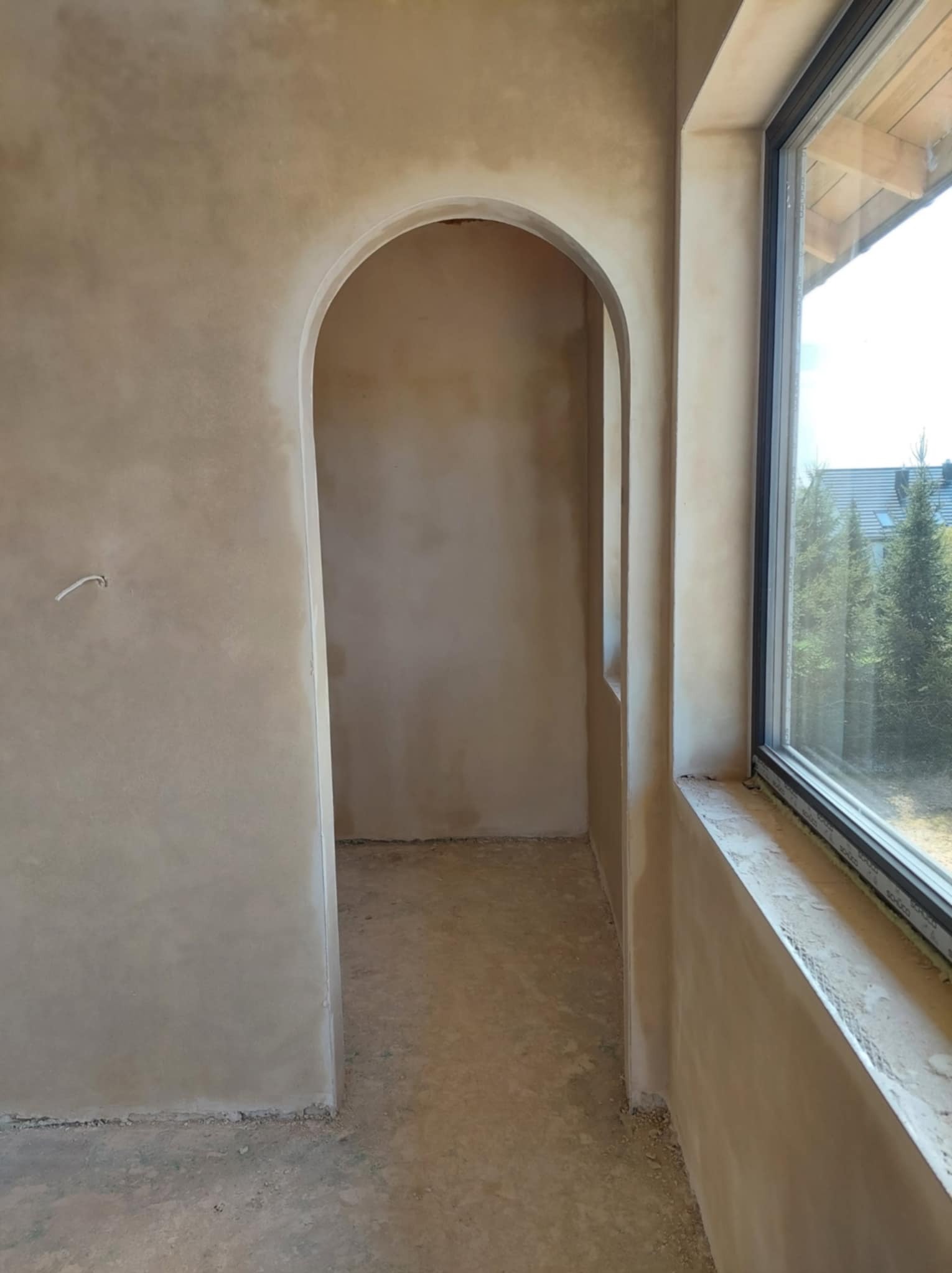 Wnętrze po szpachlowaniu: łukowate przejście, ściany o fakturze betonu, okno z widokiem na zieleń. Surowy, minimalistyczny styl wykończenia.