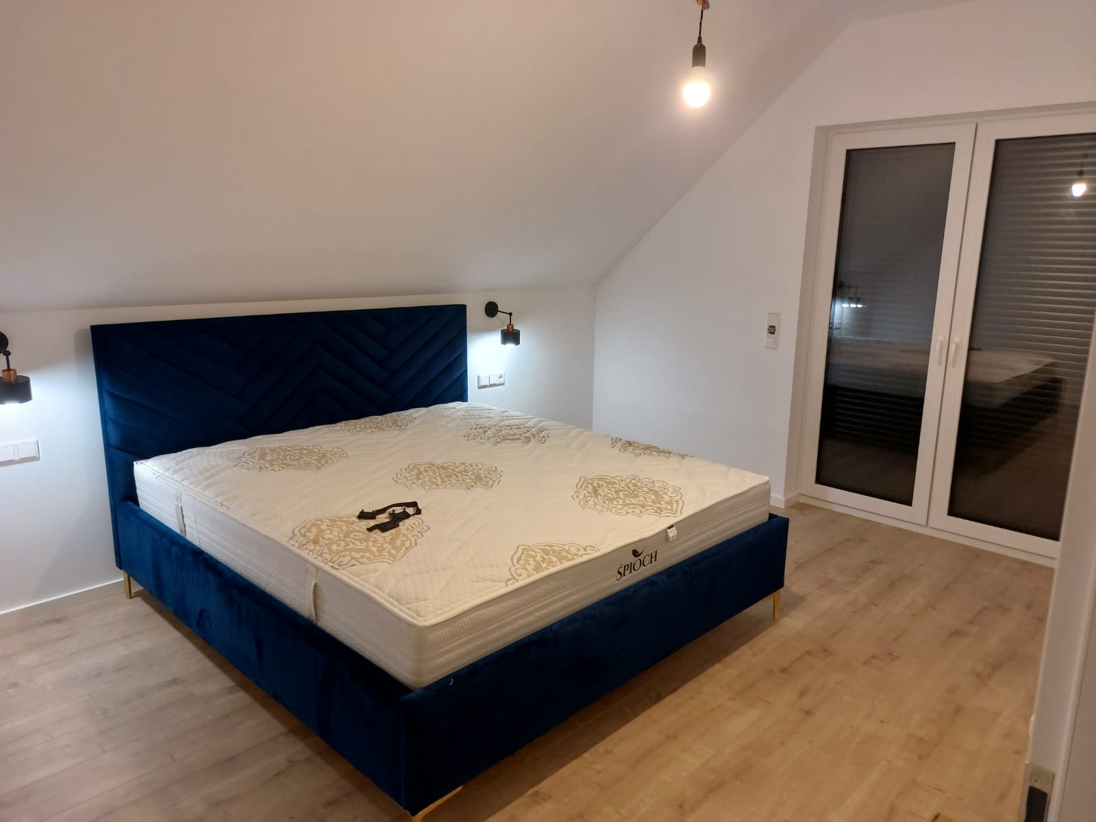Niebieskie łóżko z welurowym zagłówkiem w jodełkę, kremowy materac ze złotym wzorem w sypialni na poddaszu z drewnianą podłogą i oknem balkonowym.