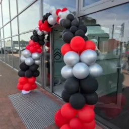 Wejście do salonu Citroena ozdobione łukiem z czarnych, srebrnych i czerwonych balonów. Widoczny fragment parkingu i elewacja budynku.