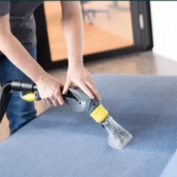 Osoba czyści jasnoniebieską tapicerkę sofy za pomocą profesjonalnego odkurzacza ekstrakcyjnego z przezroczystą końcówką, widoczne dłonie w trakcie pracy.