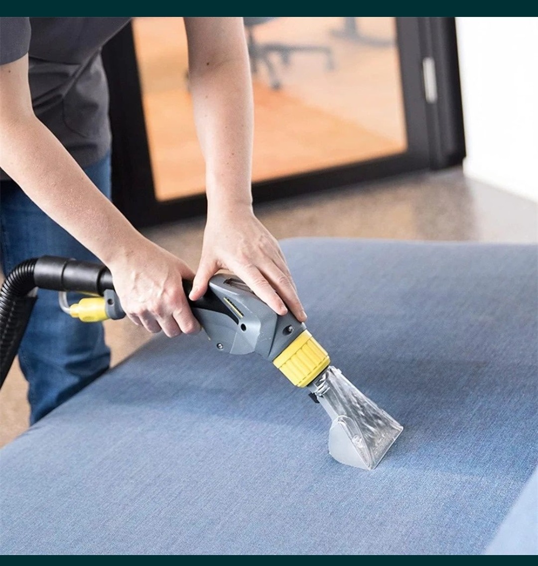 Osoba czyści jasnoniebieską tapicerkę sofy za pomocą profesjonalnego odkurzacza ekstrakcyjnego z przezroczystą końcówką, widoczne dłonie w trakcie pracy.