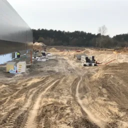 Plac budowy hali z widocznymi fundamentami, materiałami izolacyjnymi i pracownikami w odblaskowych kamizelkach na tle lasu w pochmurny dzień.