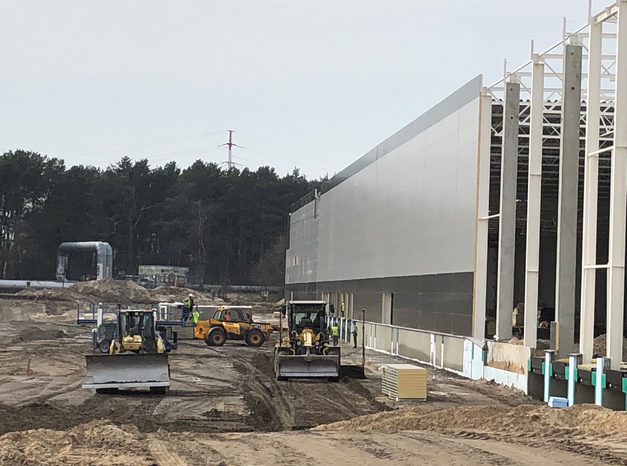 Budowa hali: widok na plac budowy z maszynami i konstrukcją stalową, w tle las i słup wysokiego napięcia.