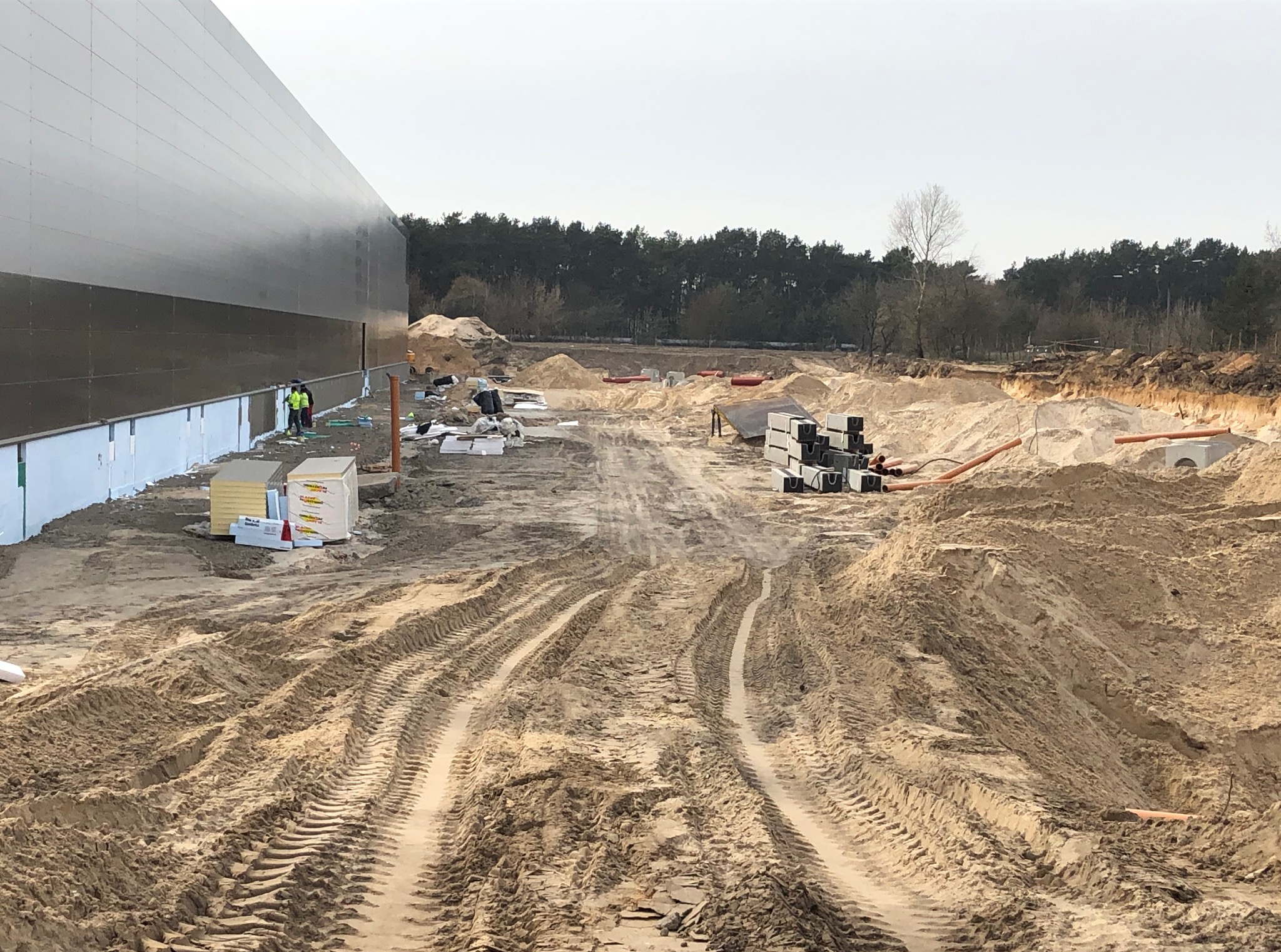 Plac budowy hali z widocznymi fundamentami, materiałami izolacyjnymi i pracownikami w odblaskowych kamizelkach na tle lasu w pochmurny dzień.