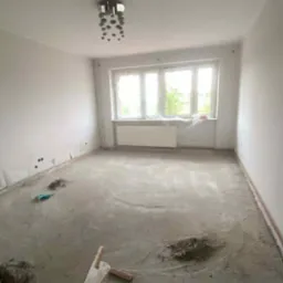 Wnętrze pokoju w trakcie prac posadzkarskich. Widoczna wylewka, narzędzia i okno z zasłoniętą folią szybą. Na suficie lampa.