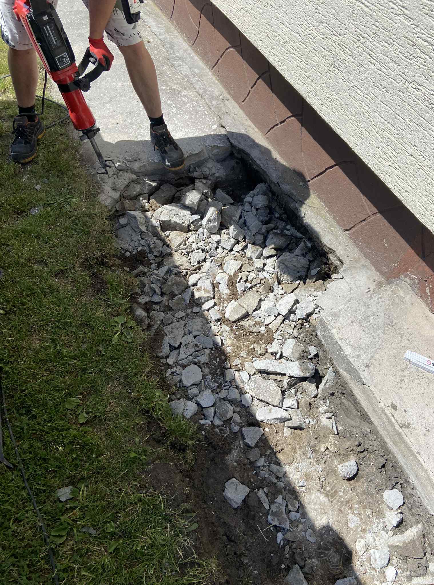 Prace rozbiórkowe przy fundamencie budynku: fragment wykopu z gruzem, pracownik z młotem pneumatycznym w rękach. Widoczna trawa i fragment ściany.