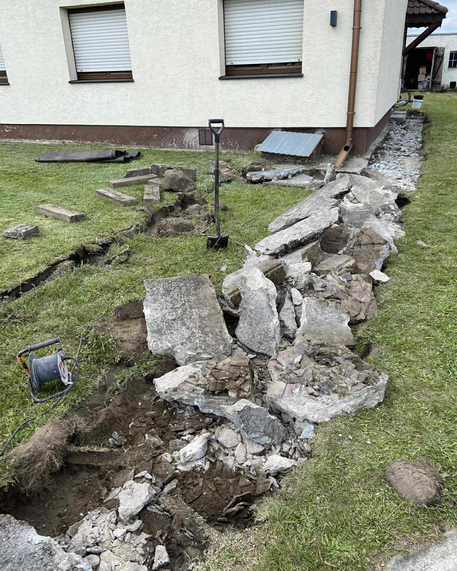 Demontaż fundamentów przy domu: gruz, wykop, łopata i przedłużacz na trawie. Fragment budynku z oknami w tle. Prace ziemne w toku.