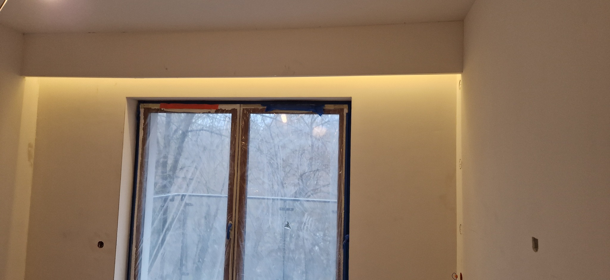 Wnęka z oświetleniem LED nad oknem balkonowym w surowym, wykończonym pokoju, z widocznymi otworami na gniazdka elektryczne w ścianie.