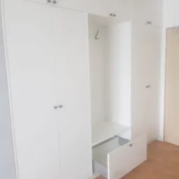 Biała szafa wnękowa z otwartą szufladą i wieszakiem, minimalistyczny design, drewniana podłoga. Widoczne detale wykończenia i metalowe uchwyty.