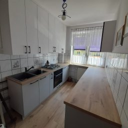 FjordWood - Meble na każdy wymiar - Jasna kuchnia z białymi szafkami, czarnymi uchwytami i blatem w odcieniu drewna. Na ścianie kwadratowe płytki z motywami. Okno z firanką w tle.