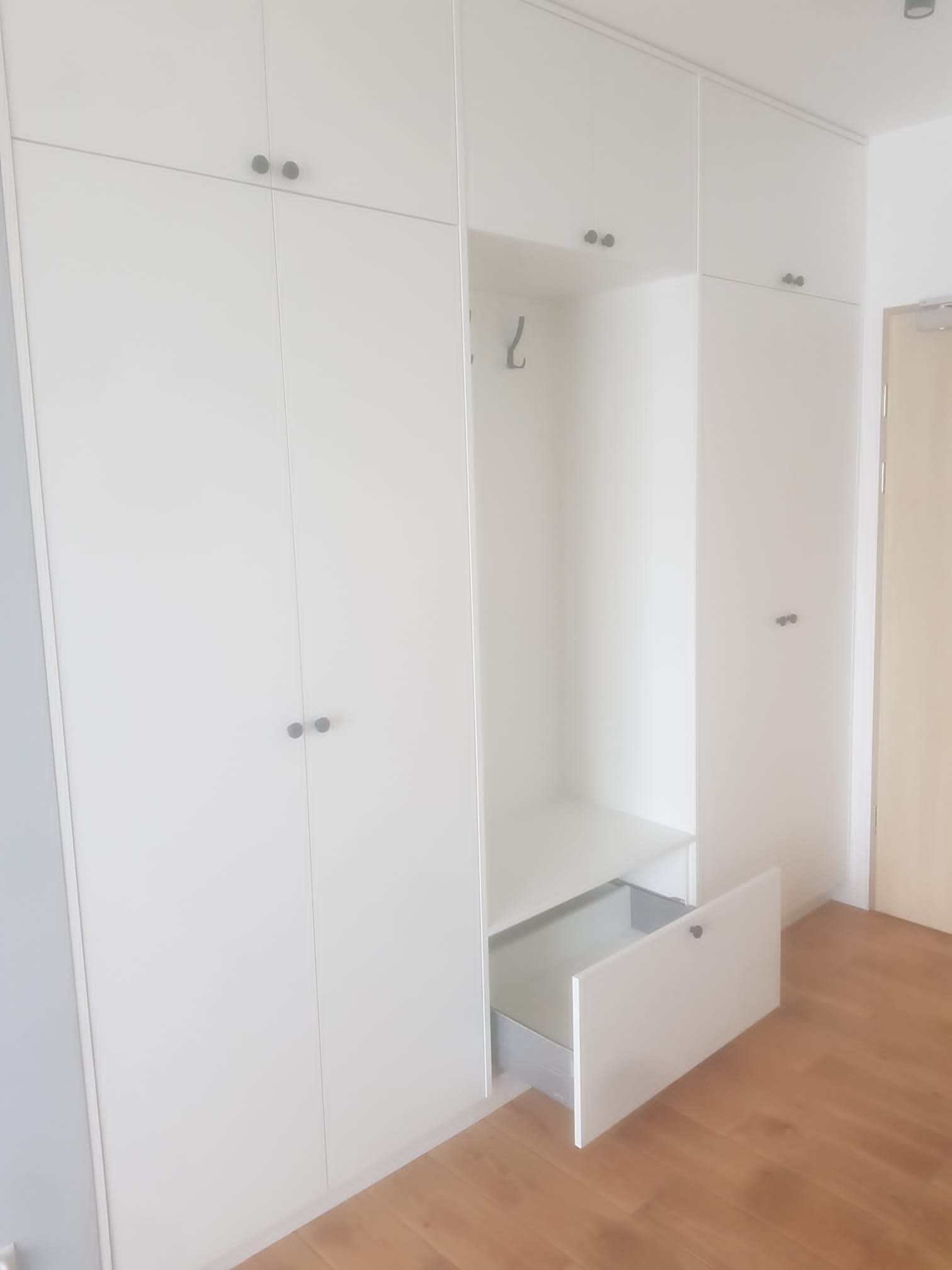 Biała szafa wnękowa z otwartą szufladą i wieszakiem, minimalistyczny design, drewniana podłoga. Widoczne detale wykończenia i metalowe uchwyty.