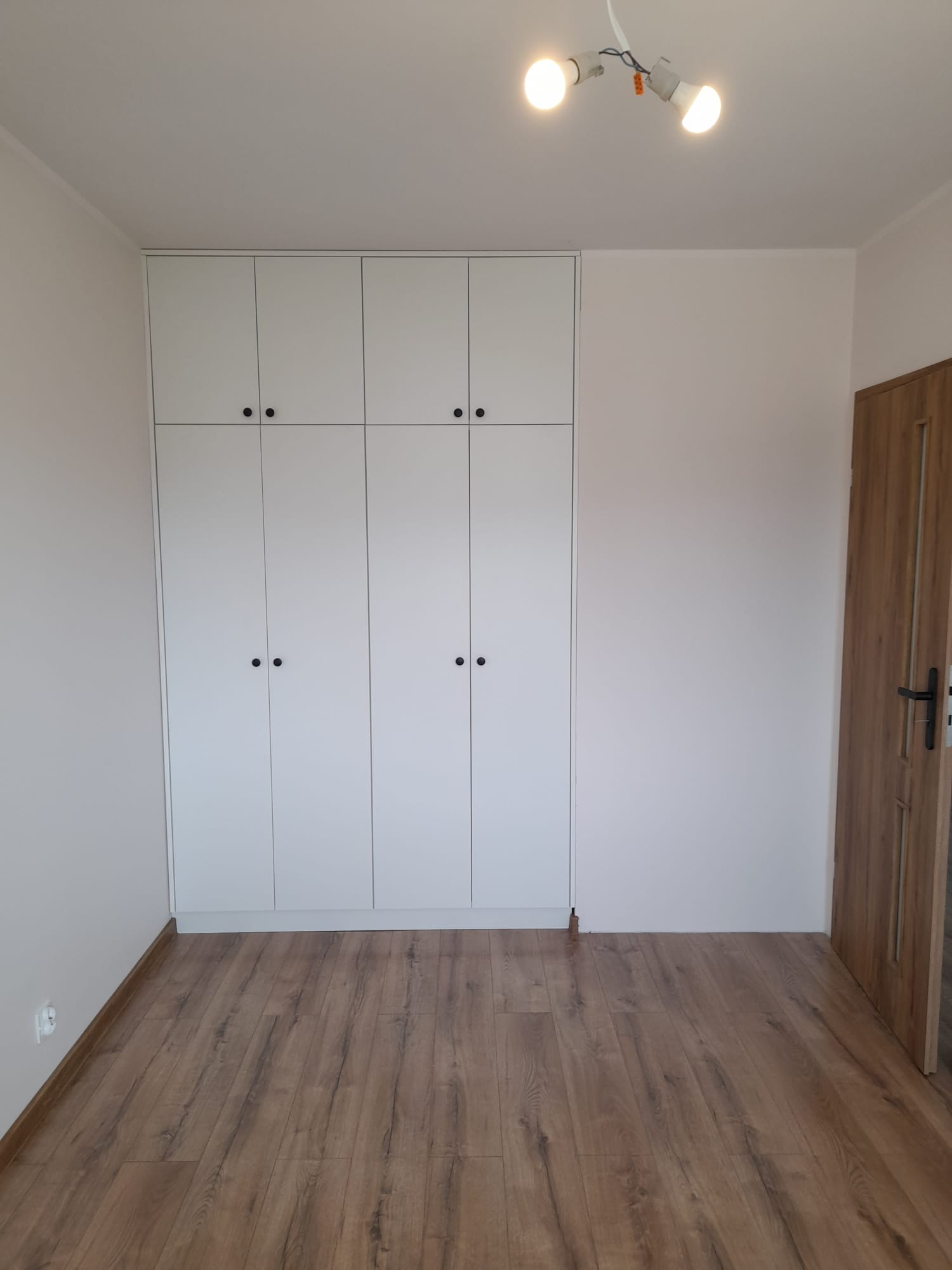 Biała szafa wnękowa z czarnymi uchwytami, dopasowana do wysokości pomieszczenia, na tle parkietu i białych ścian. Widoczne oświetlenie sufitowe.