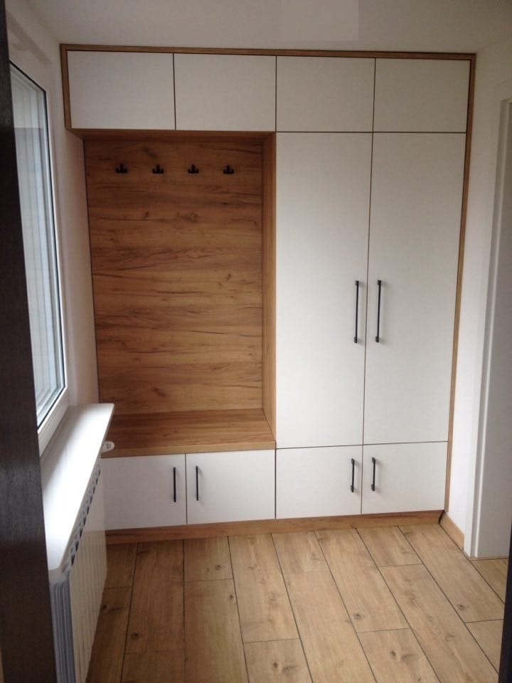 Biała szafa na wymiar z drewnianym panelem i wieszakami, dopasowana do wnęki przy oknie, na tle drewnianej podłogi, z czarnymi uchwytami.