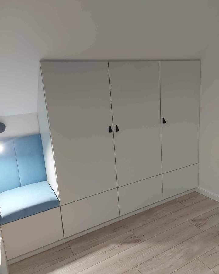 Biała szafa wnękowa z siedziskiem w kolorze błękitnym, czarne uchwyty, drewniana podłoga. Nowoczesny design, dopasowanie do skosu.