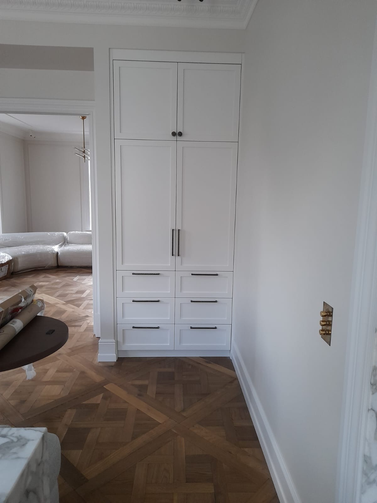 Biała szafa wnękowa z ciemnymi uchwytami, wkomponowana w ścianę, na tle parkietu w jodełkę i częściowo zasłoniętego mebla w salonie.