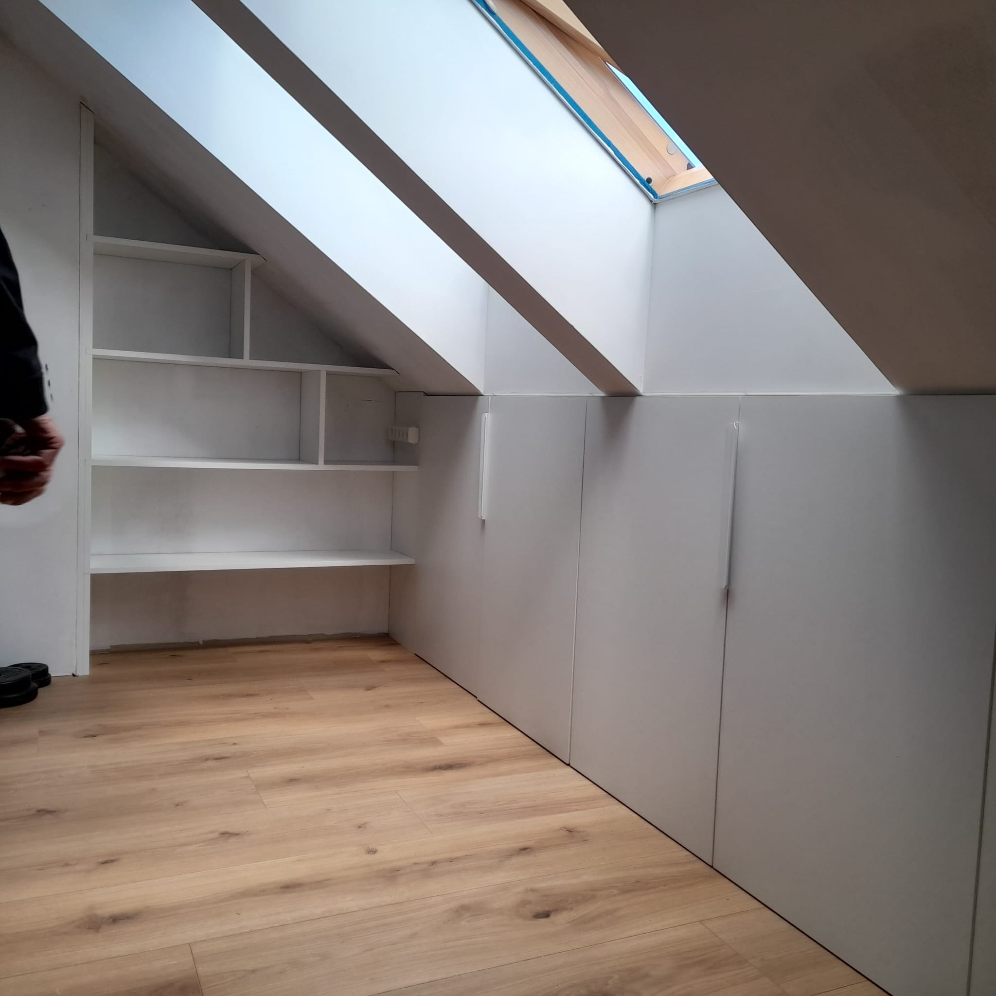 Biała zabudowa wnęki na poddaszu ze skośnym oknem dachowym. Po lewej stronie otwarte półki, po prawej szafki z podłużnymi uchwytami.