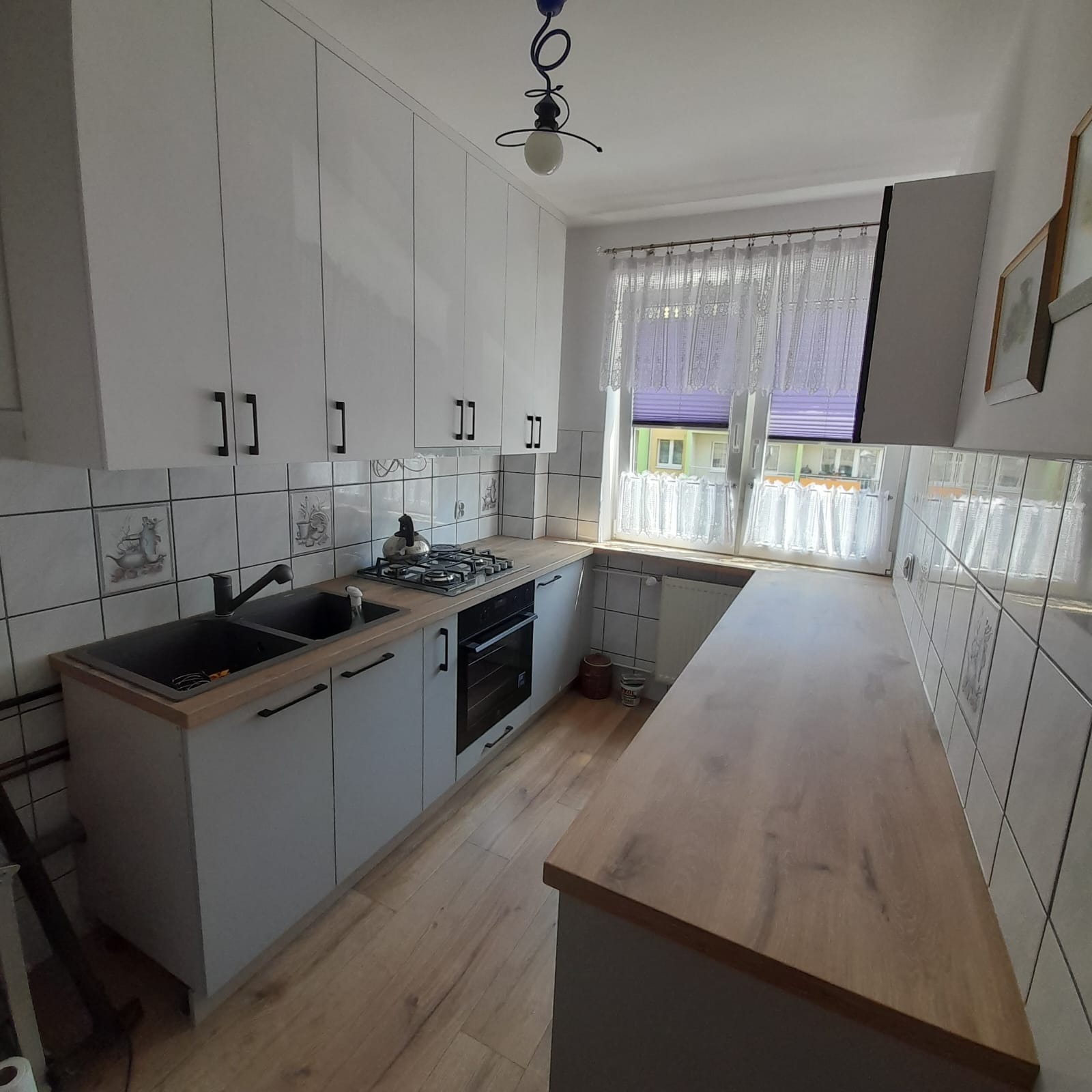 Jasna kuchnia z białymi szafkami, czarnymi uchwytami i blatem w odcieniu drewna. Na ścianie kwadratowe płytki z motywami. Okno z firanką w tle.