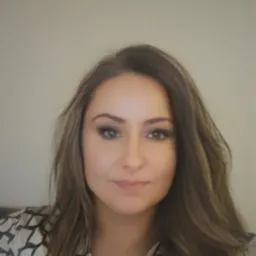 Portret kobiety z długimi, brązowymi włosami i subtelnym makijażem, uśmiechającej się delikatnie. Widoczna bluzka we wzory i jasne tło.