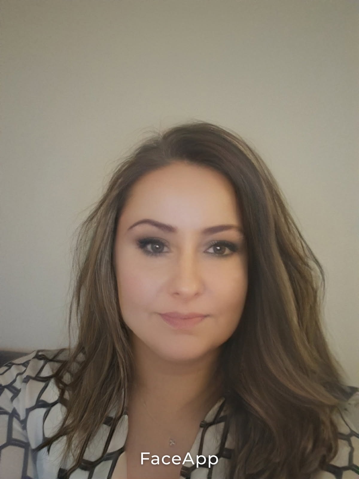 Portret kobiety z długimi, brązowymi włosami i subtelnym makijażem, uśmiechającej się delikatnie. Widoczna bluzka we wzory i jasne tło.