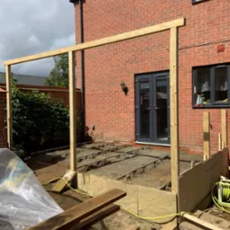 Konstrukcja drewniana tarasu w trakcie budowy przy domu z cegły. Widoczne deski i elementy montażowe. Etap przygotowania podłoża i wznoszenia ramy nośnej.