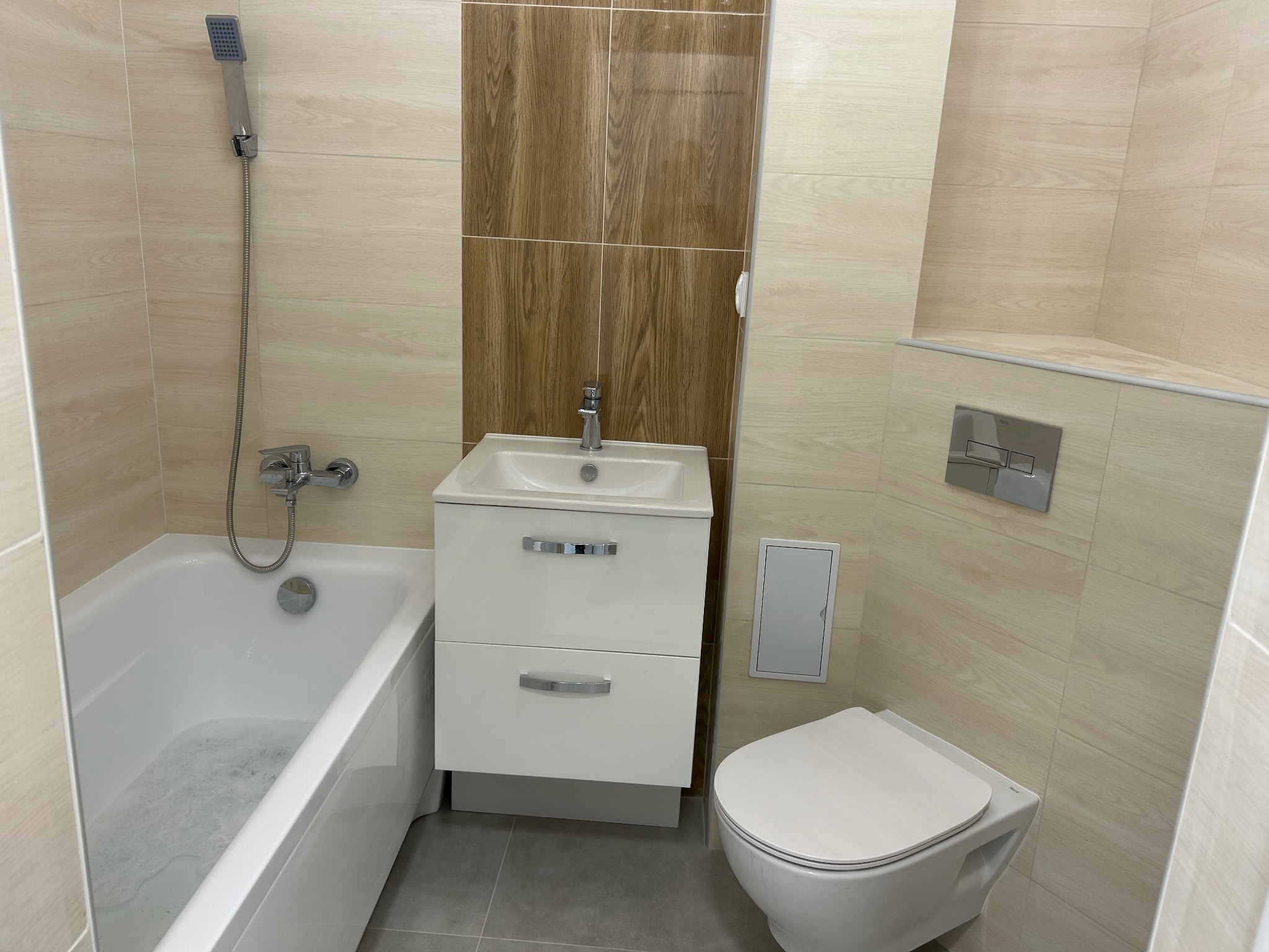 Jasna łazienka po remoncie: beżowe płytki imitujące drewno, biała wanna z prysznicem, umywalka z szafką i toaleta. Kontrastowe, ciemniejsze płytki za umywalką.